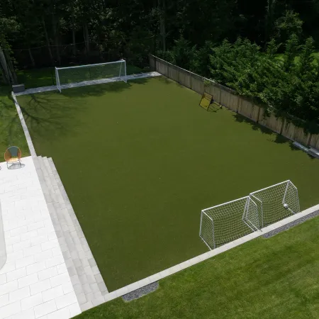 A backyard featuring a small soccer field with two goals, patio with pool, chairs, and surrounding greenery.