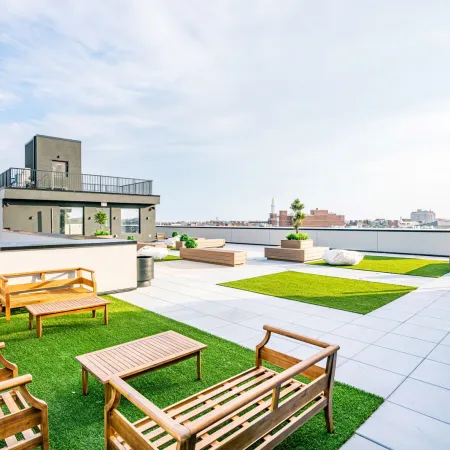 Spacious rooftop patio with wooden benches and tables on green artificial turf under blue sky in urban setting