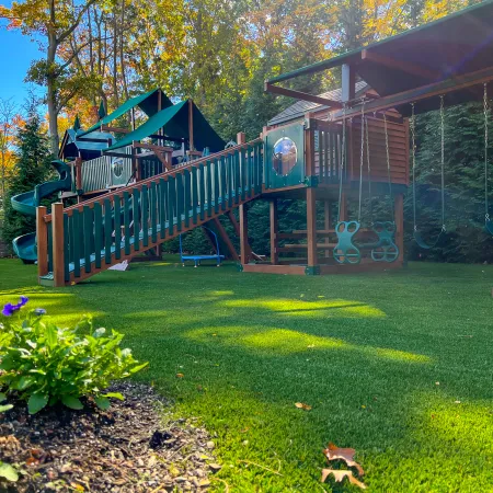 Children's wooden playground with slide, swings, and climbing structure surrounded by green lawn and trees.