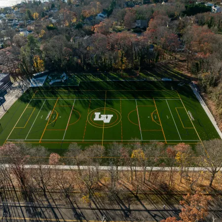 Aerial view of a green sports field with white and orange markings and LV logo surrounded by autumn trees and buildings.