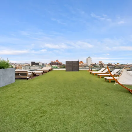 Rooftop lounge area with artificial grass, hammocks, lounge chairs, and city skyline under a blue sky.