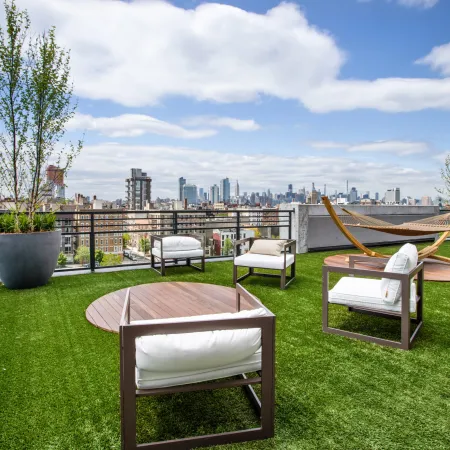 Modern rooftop patio with cushioned chairs, hammock, potted trees, and city skyline under a blue sky.