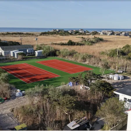 Aerial view of two red tennis courts surrounded by greenery near a coastal residential area under clear blue sky.