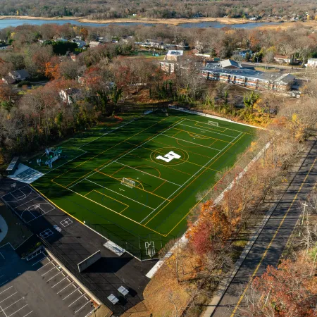Aerial view of a green sports field with soccer goals and a large H logo surrounded by autumn trees and buildings.