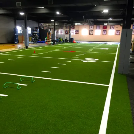 Indoor sports training facility with artificial turf, agility hurdles, cones, and gym equipment in a spacious room.
