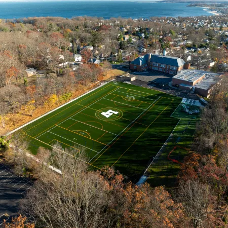 Aerial view of a green sports field with markings and a large H logo, surrounded by autumn trees and buildings near a coastline.