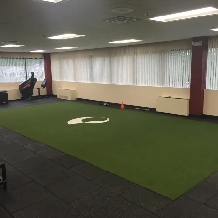 Empty indoor gym area with green turf, exercise equipment, and large windows with vertical blinds.