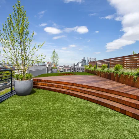 Modern rooftop terrace with curved wooden deck, large potted trees, green grass, and city skyline under a blue sky.
