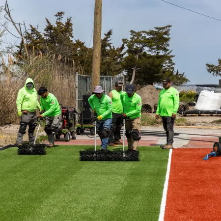Team of workers in bright green shirts installing artificial turf on a sports field during daytime outdoors.