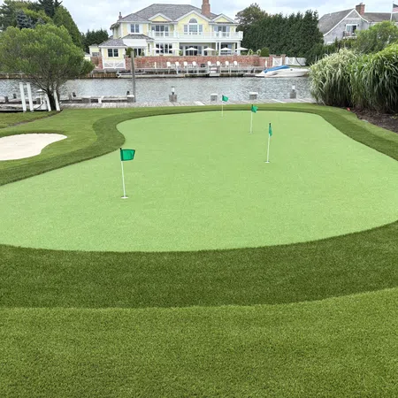 Golf putting green with multiple flags next to a waterfront house and dock under a cloudy sky.