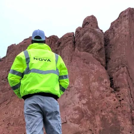 Person in neon green reflective jacket and cap facing large red rock formations under overcast sky
