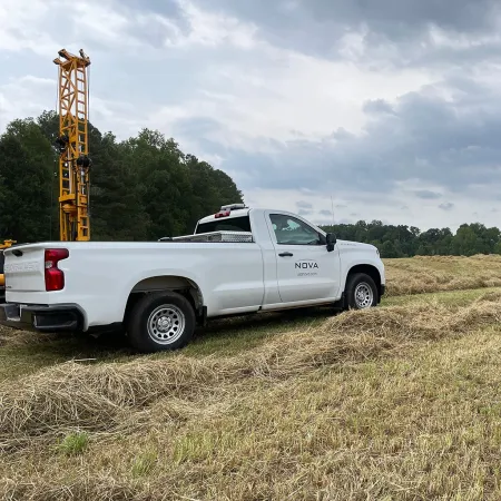 White pickup truck with NOVA logo parked on grassy field near drilling equipment under cloudy sky