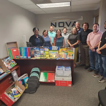 Group of people standing behind a desk filled with school supplies including notebooks, backpacks, and writing tools