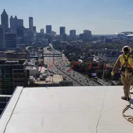 Construction worker with safety harness on rooftop overlooking a busy highway and city skyline under clear sky.