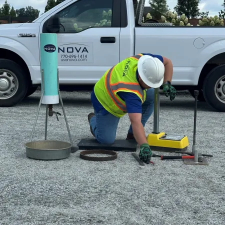 Construction worker in safety gear testing gravel compaction near Nova company truck outdoors.