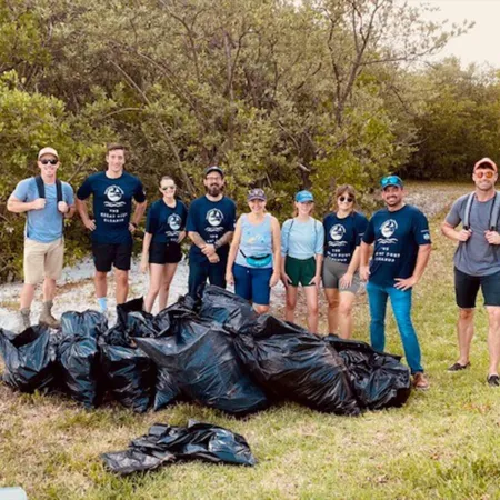 Group of volunteers posing with collected trash bags during an outdoor cleanup event in a grassy area.