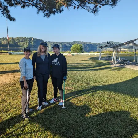 Three women standing on a grassy field with golf clubs, smiling under clear blue sky with trees and picnic shelters.
