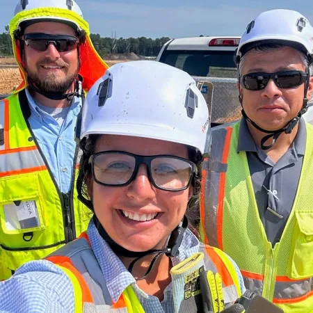 Three construction workers wearing hard hats and high-visibility vests smiling at a construction site with vehicles.