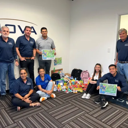 Group of nine people in NOVA shirts with donated school supplies displayed on floor inside office.