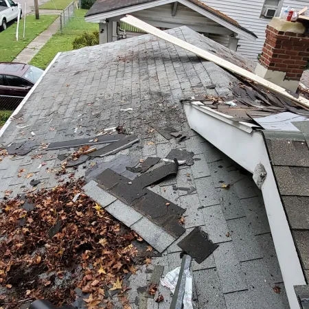 Damaged roof with missing shingles, debris, and fallen leaves near a white house and brick chimney.