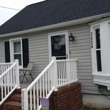 Small gray house with white railings, brick steps, black shutters, and a front porch with two chairs.