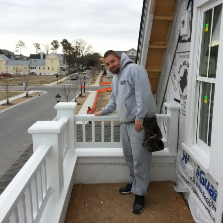 Man in gray hoodie and sweatpants working on a white balcony railing of a house under construction.