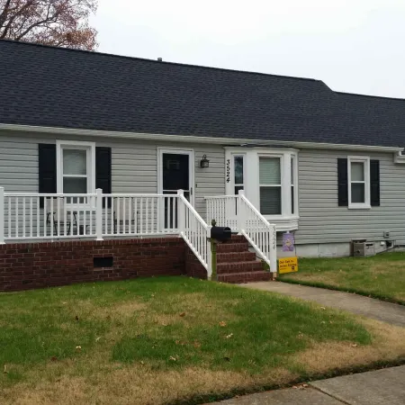 Single-story gray house with black shutters, white porch railing, and green lawn on a residential street.