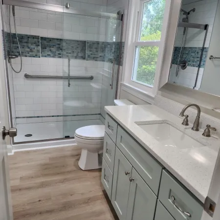 Modern bathroom with glass shower enclosure, mosaic tile accent, white vanity, and wood-look flooring under bright natural light