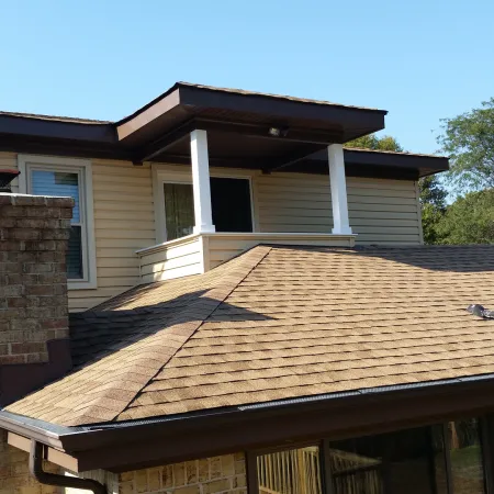 Two-story house with brown shingle roof, beige siding, brick chimney, and white pillars on balcony under clear sky