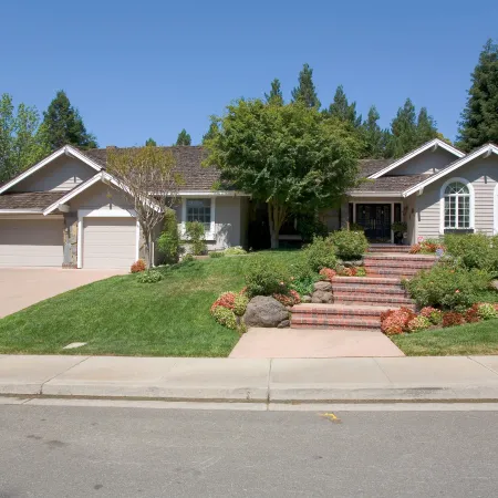 Single-story house with brick steps, manicured lawn, and mature trees under a clear blue sky.