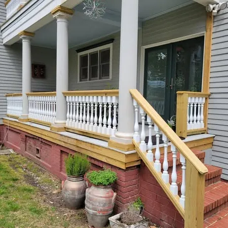 Front porch with white balusters, wooden railings, brick foundation, and potted plants in front yard