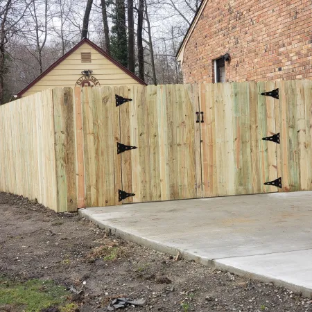 New wooden privacy fence with double gate installed next to brick house and concrete driveway.