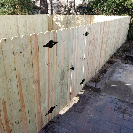 Newly installed wooden privacy fence with gate and black metal hinges surrounds patio area with stone and concrete flooring.