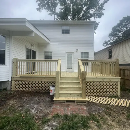 Newly built wooden deck with railing and steps attached to white two-story house with backyard area.