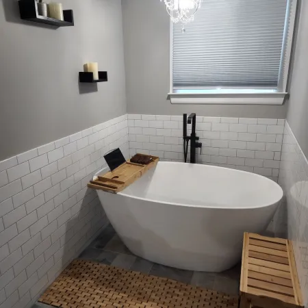 Modern bathroom with freestanding white tub, wooden bath tray, woven floor mat, and decorative shelves with plants and candles.
