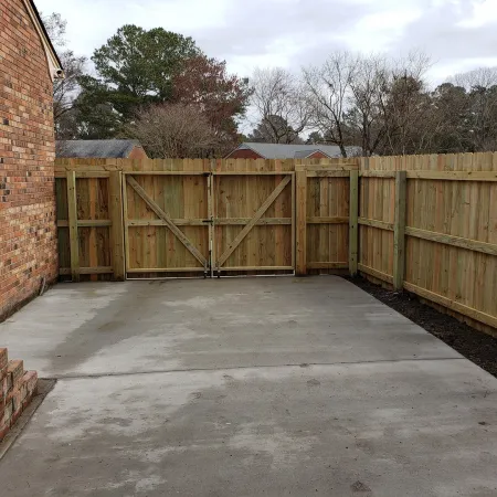 Concrete driveway area bordered by wooden fence and brick house wall with steps leading to the door.