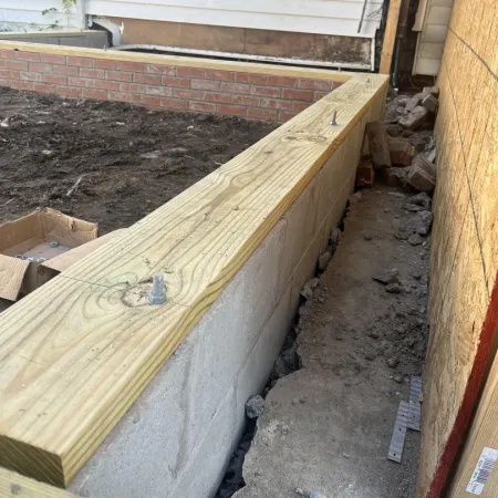 Foundation wall with wooden sill plate installed on concrete blocks next to house under construction.