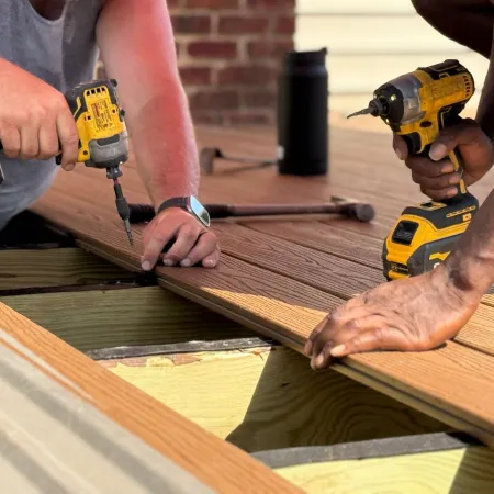 Two workers installing wooden deck planks using cordless power drills on a sunny day.