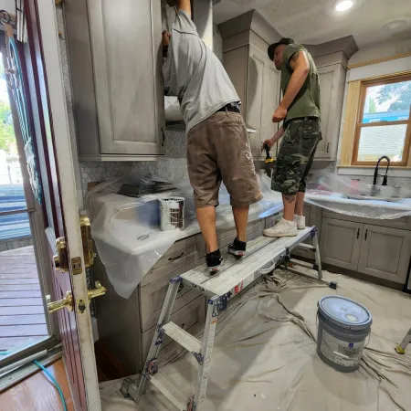 Two workers installing gray kitchen cabinets using a folding ladder in a home renovation setup