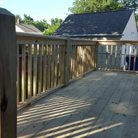 Sunlit wooden deck with vertical railing and shadows, attached to a house with neighboring roofs visible.