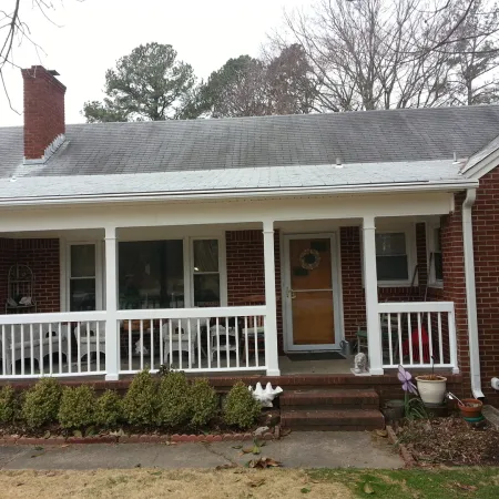 Front view of a brick house with white porch railing, wooden door, shrubs, and overcast sky.