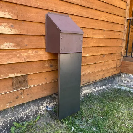 Black metal vent pipe extending from a wooden deck to grassy ground with a stone wall nearby.