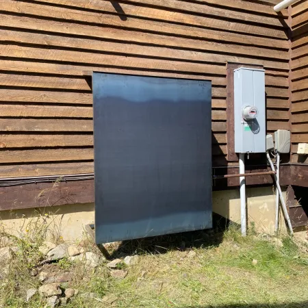 Metal panel mounted on the side of a wooden building next to an electrical box in sunny outdoor setting.