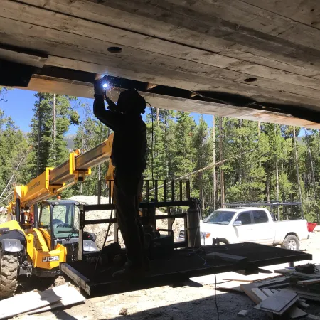 Welder working on a construction site with a yellow JCB telehandler and pickup truck in a forested area