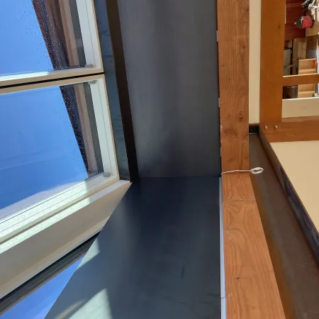 Modern interior corner with large windows, natural wood beams, and sunlight casting shadows on dark surfaces