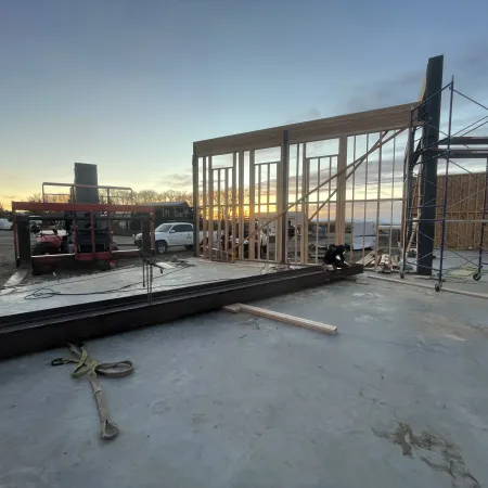 Construction site at sunset with wooden framing, scaffolding, machinery, and workers assembling a building.