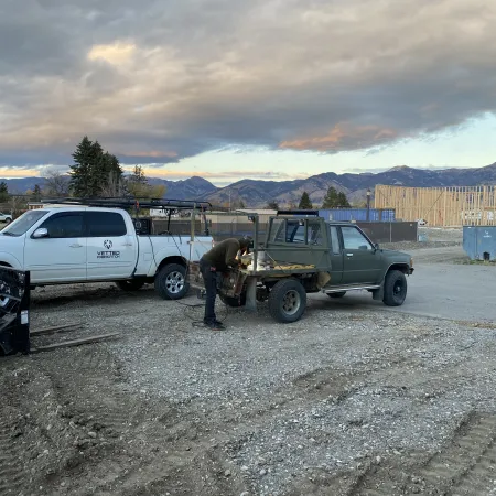 Worker repairs old pickup truck at construction site with mountains and cloudy sky in the background.