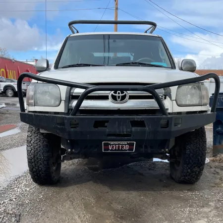Front view of a white Toyota truck with off-road bumper and large tires parked on gravel near trash bins.