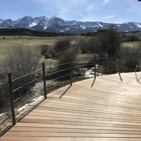 Curved wooden deck with metal railing overlooking a green field and snow-capped mountains under a blue sky