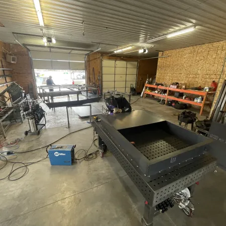 Welder works on large metal table in spacious workshop filled with tools and equipment under fluorescent lights.